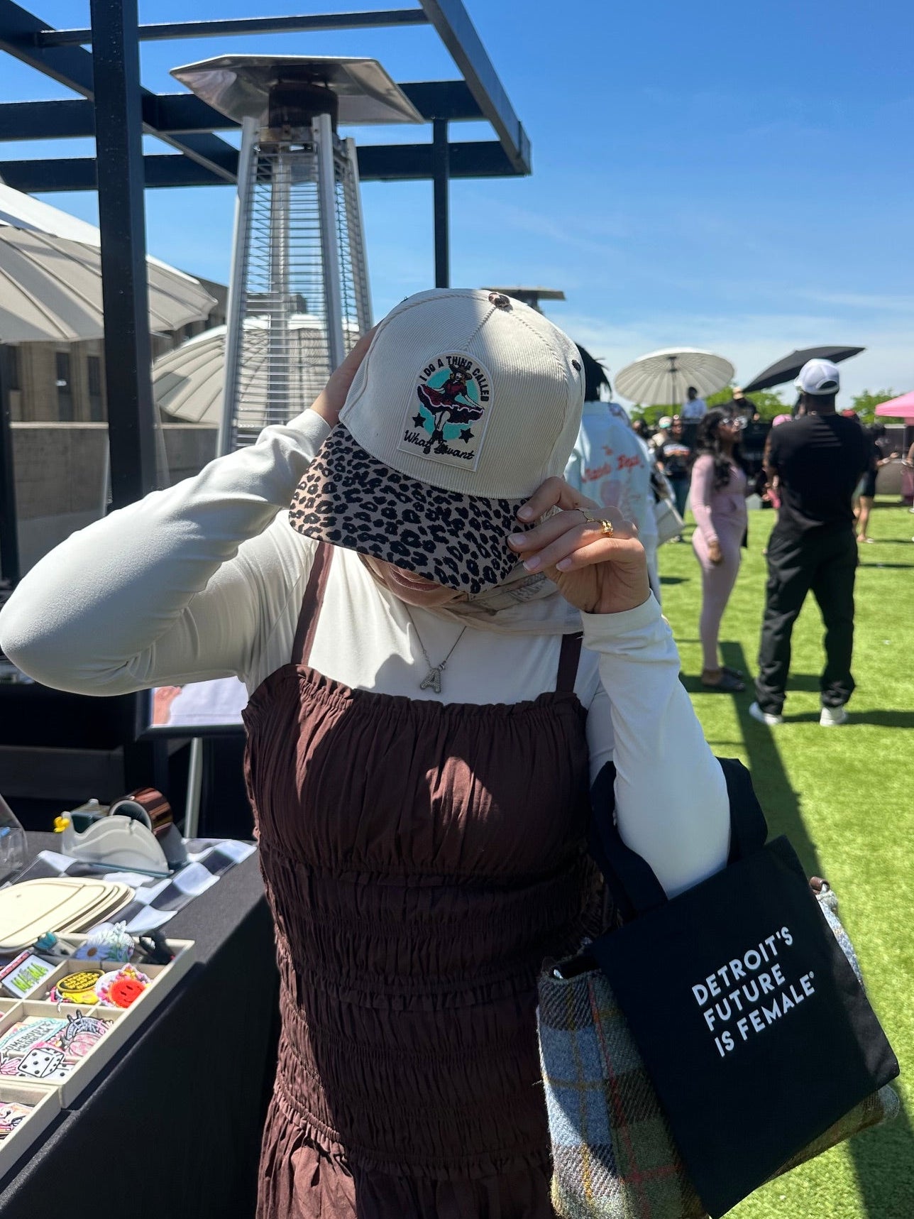 Person wearing a cheetah print cap with a logo and patterned hijab, standing outdoors on a sunny day.